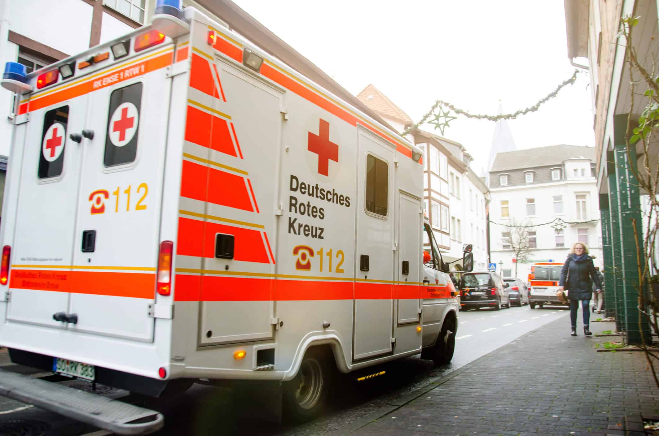 A Deutsches Rotes Kreuz (German Red Cross) ambulance is parked on a street in a European city; a person walks on the sidewalk nearby. Beekeeper